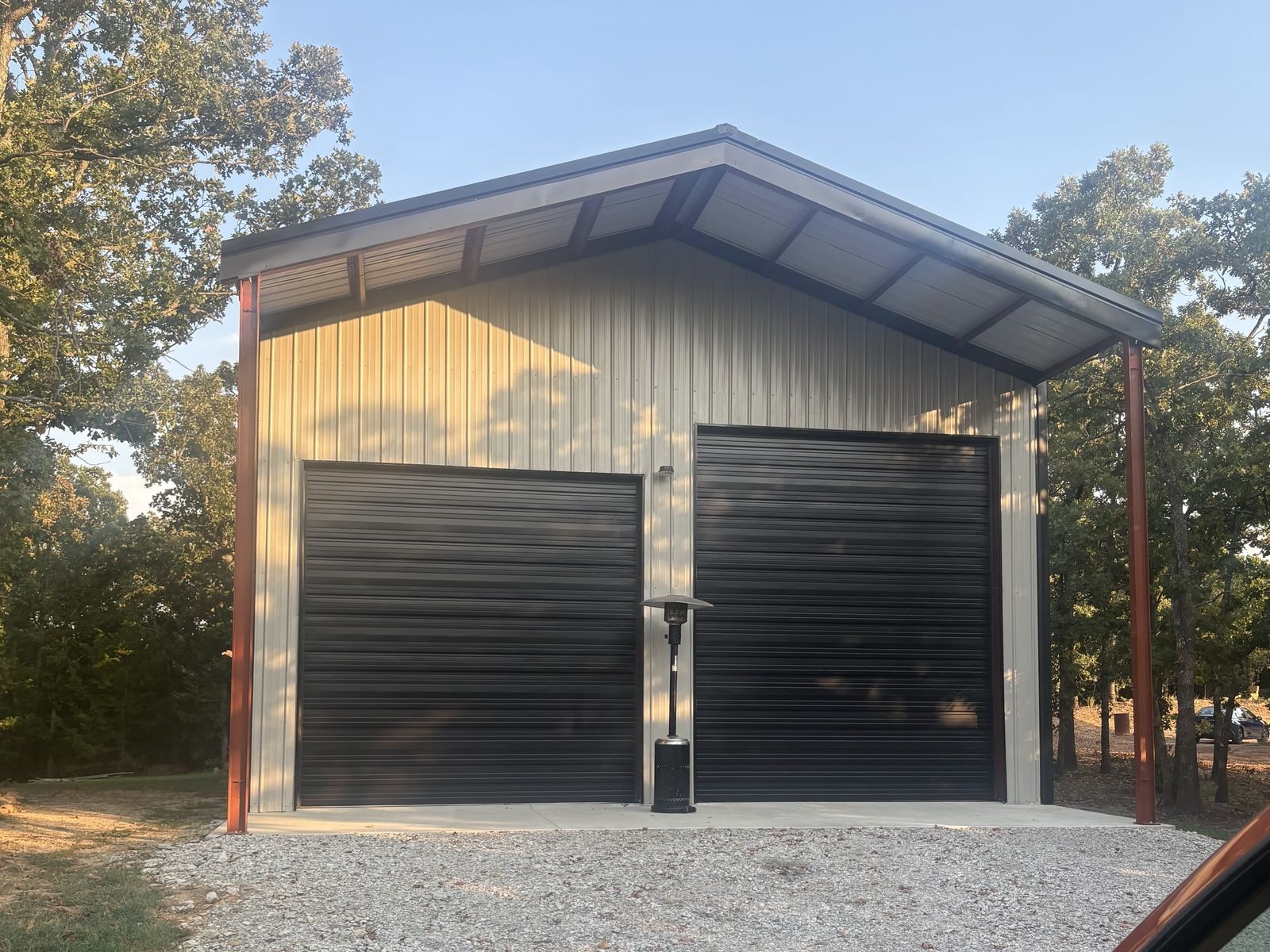 Tan metal garage with two black roll up doors on a gravel drive